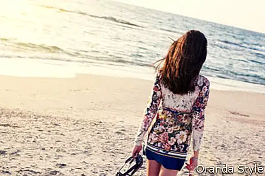 junge und schöne Frau im bunten Kleid, das auf den Strand nahe dem Ozean geht und weit weg den Sonnenuntergang betrachtet
