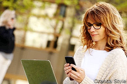 Портрет на заета жена от продажбите, седнала в бизнес парка и използвайки своя лаптоп, докато пише текст на мобилен телефон