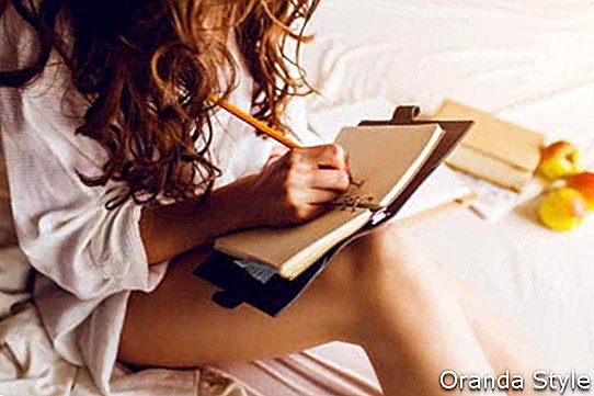 Menutup imej lembut gaya hidup wanita cantik muda yang duduk di atas katilnya yang selesa dan membuat nota ke buku hariannya
