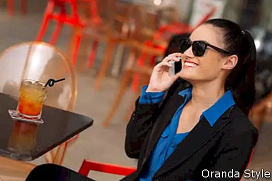 Весела бизнес жена в слънчеви очила, седнала в кафенето и разговаряща по телефона