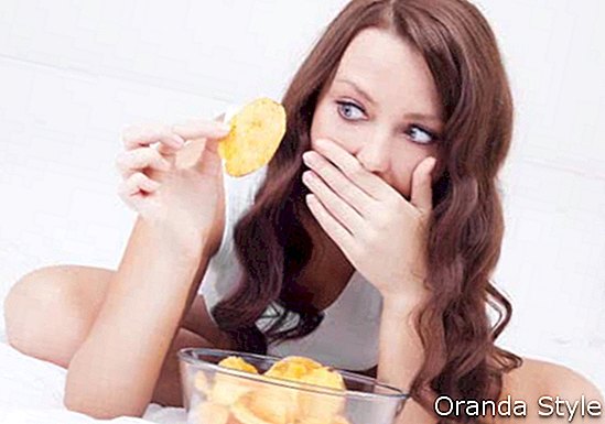 вродлива жінка їсть картопляні чіпси в ліжку вдома і боїться набрати вагу