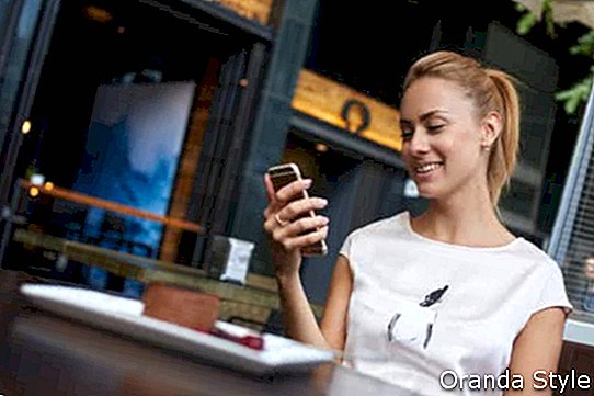 Junge glückliche Frau, die gute Nachrichten an ihrem Handy beim Sitzen im modernen Kaffeestubeinnenraum liest