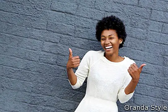 Porträt der lächelnden jungen Afroamerikanerfrau, die Daumen gestikuliert, up Zeichen mit beiden ihren Händen gegen graue Wand