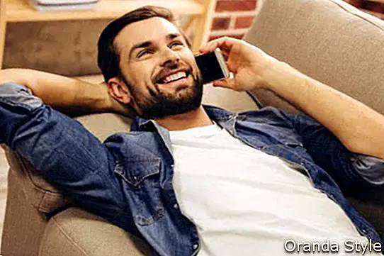 Красивият мъж в дънкови дрехи говори по мобилния телефон и се усмихва, докато лежи на дивана