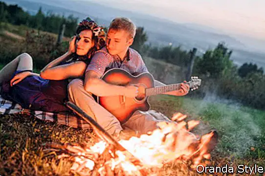 Glückliche junge Paare, die durch Feuer mit Gitarre sitzen