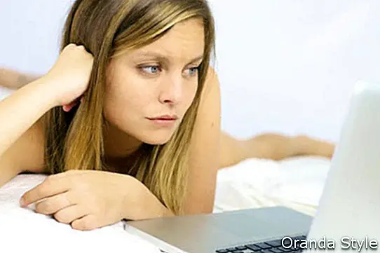 Тужна жена изгледа за рачунаром како лежи у кревету