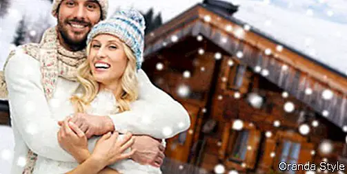 lächelnder Mann und Frau in den Hüten und im Schal, die über hölzernem Landhaus- und Schneehintergrund umarmen