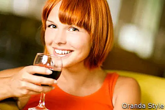 Jauna skaisti smaidīga sieviete sarkanā vestē sēž atzveltnes krēslā un viņai ir kontrole pār glāzi ar vīnu