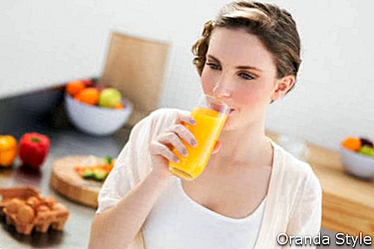 Burvīga brunete, sieviete dzer glāzi apelsīnu sulas, stāvot savā virtuvē mājās
