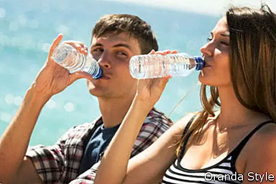 Pasangan muda yang dahaga menikmati sebotol air di tepi pantai pada hari yang cerah