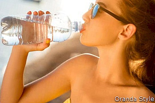 Mlada žena koja pije pjenušavu vodu iz prozirne boce na plaži na zalazak sunca