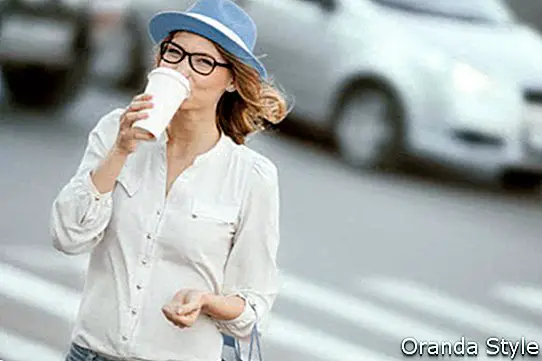 סטודנטית אופנתית צעירה ומאושרת ששותה קפה חם לקחת ממנה וחוצה דרך על רקע עירוני