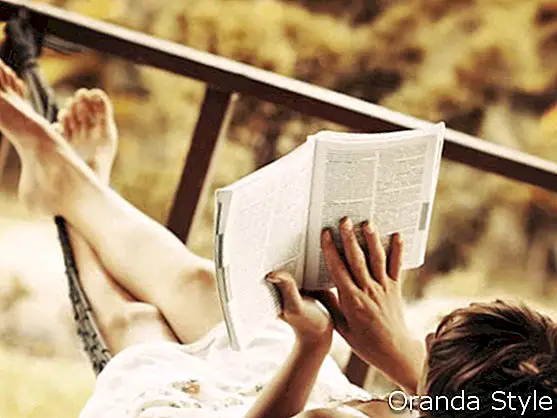 Wanita berbaring di buaian di taman dan menikmati bacaan buku