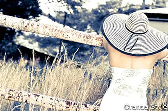 гърба на жена, облечена в голяма шапка, облегнала се на ръждясала ограда