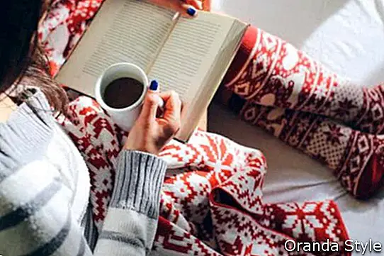 Meitene gultā ar kafijas tasi, lasot grāmatu