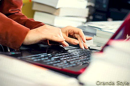 Frau, die auf der Laptoptastatur umgeben durch Bücher und Dateien schreibt