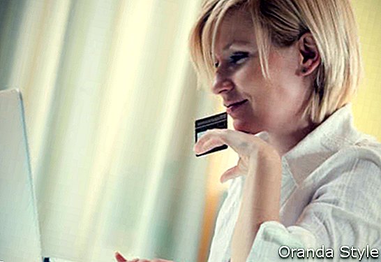 Млада насмејана жена код куће користећи рачунар за интернет куповину држећи у руци кредитну картицу