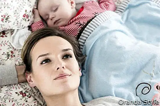 una joven madre acostada en una cama junto a su bebé dormido