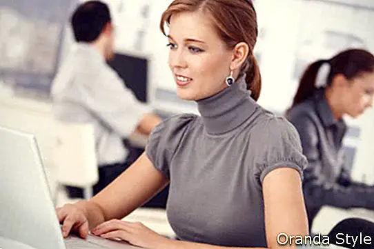 אישה צעירה עובדת במשרד, יושבת ליד שולחן העבודה באמצעות מחשב נייד