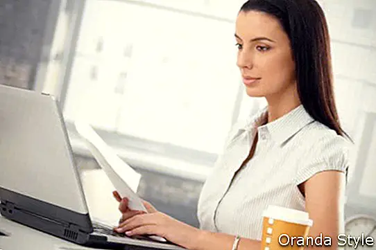 कार्यालय में डेस्क पर बैठी आकर्षक महिला लैपटॉप कंप्यूटर होल्डिंग डॉक्यूमेंट के साथ टेकअवे कॉफी ले रही है