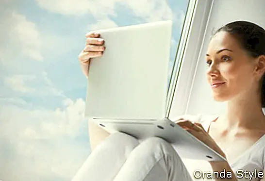 Жена која сједи поред прозора и користи лаптоп