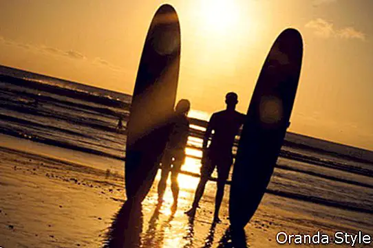 Surferpaare im Schattenbild, das lange Brandungsbretter bei Sonnenuntergang hält
