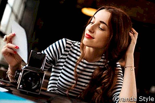 Jaunas sievietes fotogrāfs, kurš skatās uz izdrukātajām fotogrāfijām ar vecu 6x6 kadru kameru, sēžot kafejnīcā ar bēniņu dizaina interjeru