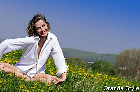 Atractive Frau mit dem blonden Haar und einer weißen Bluse, zum eines reichen blühenden Frühlingswiesenyogas und -entspannungsübungen gegen einen blauen Himmel zu machen