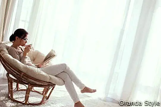 Mujer joven en su casa sentada en una silla moderna frente a la ventana, relajarse en su sala de estar leyendo un libro y tomando café o té