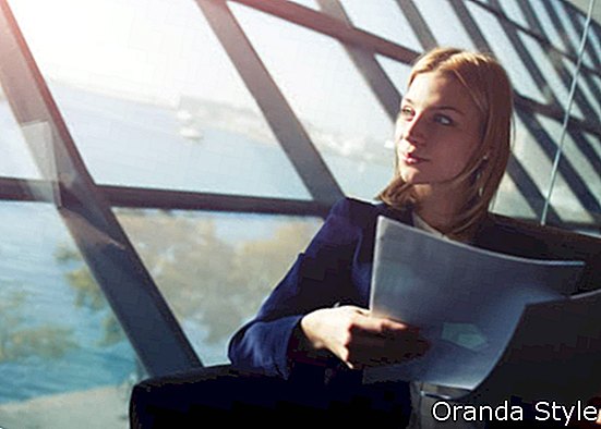 Potret wanita elegan muda duduk di dalam kertas kerja dalaman pejabat moden dan menatap keluar dari tingkap +