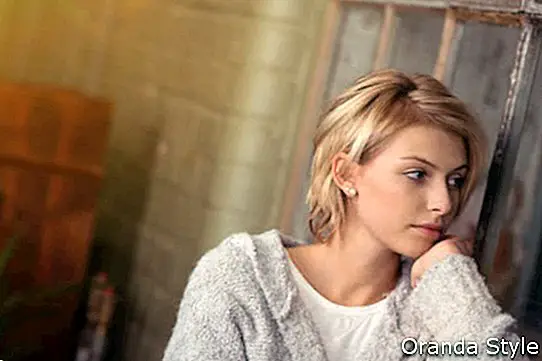 Mlada žena sjedi dok sanja ili čeka da netko pogleda kroz prozor u dnevnoj sobi s ozbiljnim izrazom lica
