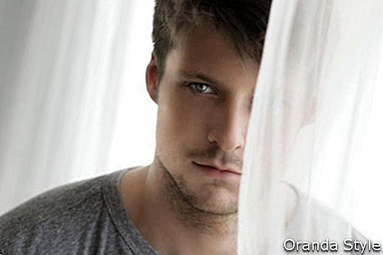 Stående av en ung man nära fönstret med den genomskinliga gardinen som döljer hälften av hans ansikte