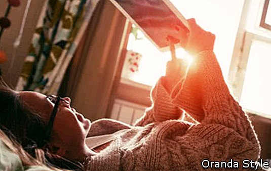 naine, kes lamab voodis koos digitaalse tahvelarvutiga, mida hommikuse päikesevalguse käes sõrmega puudutada