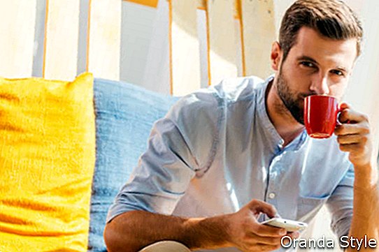 Hübscher junger Mann, der Handy hält und Kaffee beim Sitzen in der Ruhezone des Büros 2 trinkt