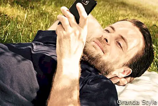 איש מסתכל בטלפון נייד בזמן הנחת דשא בפארק במהלך יום קיץ שטוף שמש