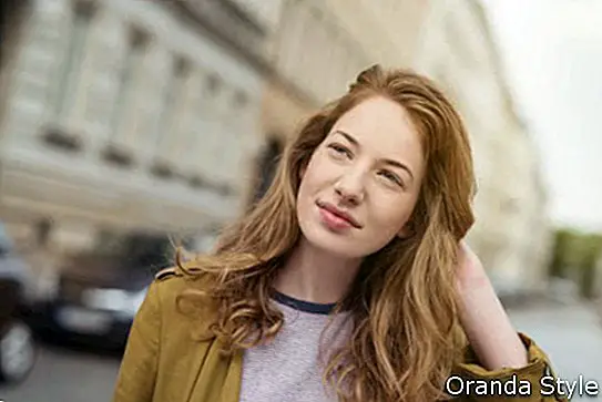 Ελκυστική νεαρή γυναίκα που στέκεται σε ένα αστικό δρόμο σκέψης βαθιά κοιτάζοντας προς τα έξω με μια μακρινή έκφραση και το χέρι της στα μαλλιά της